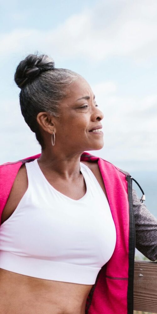 A woman in athletic wear leans on a wooden fence by the ocean, holding a yoga mat and gazing peacefully into the distance.