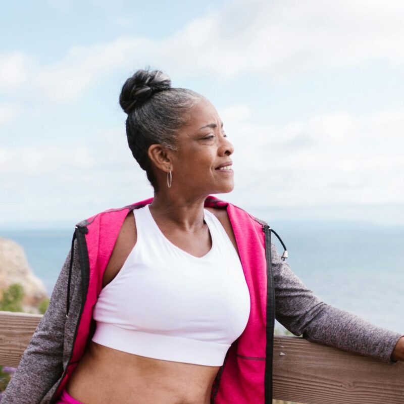 A woman in athletic wear leans on a wooden fence by the ocean, holding a yoga mat and gazing peacefully into the distance.