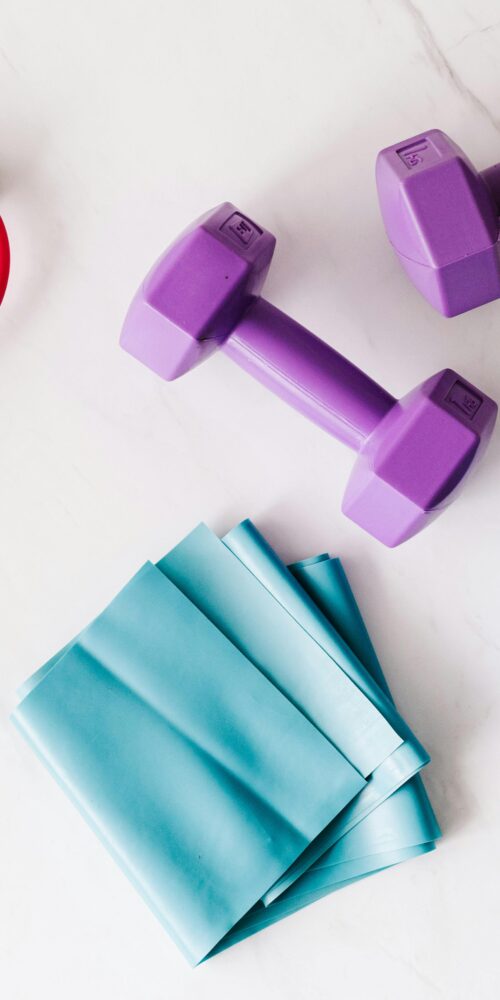 Fitness equipment on a white marble surface, including a resistance band with foam handles, a pair of dumbbells, and a blue double lacrosse massage ball.