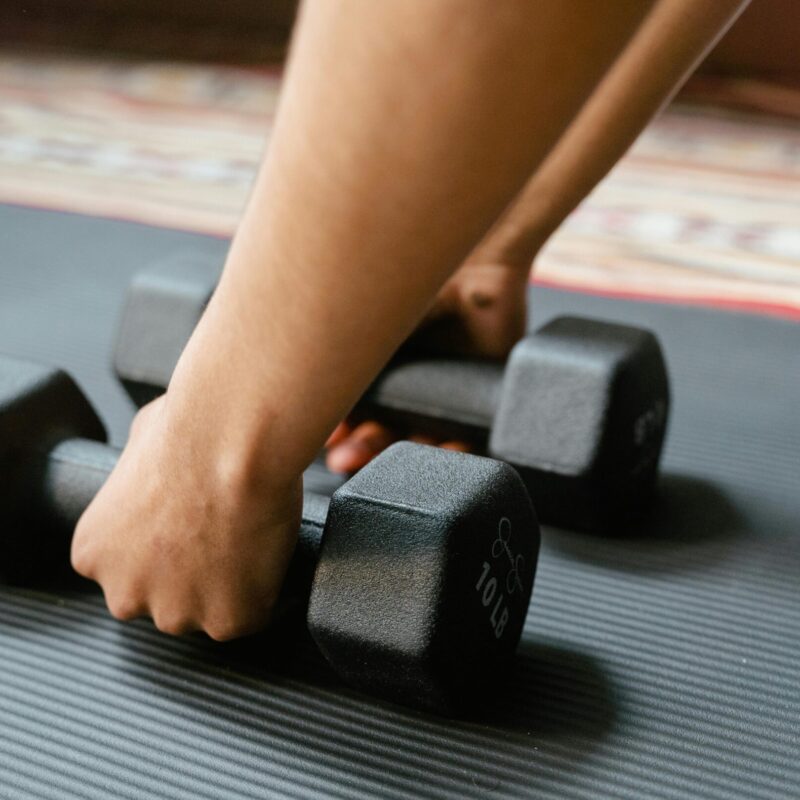 Close-up of a person picking up two black 10-pound dumbbells from a textured exercise mat.