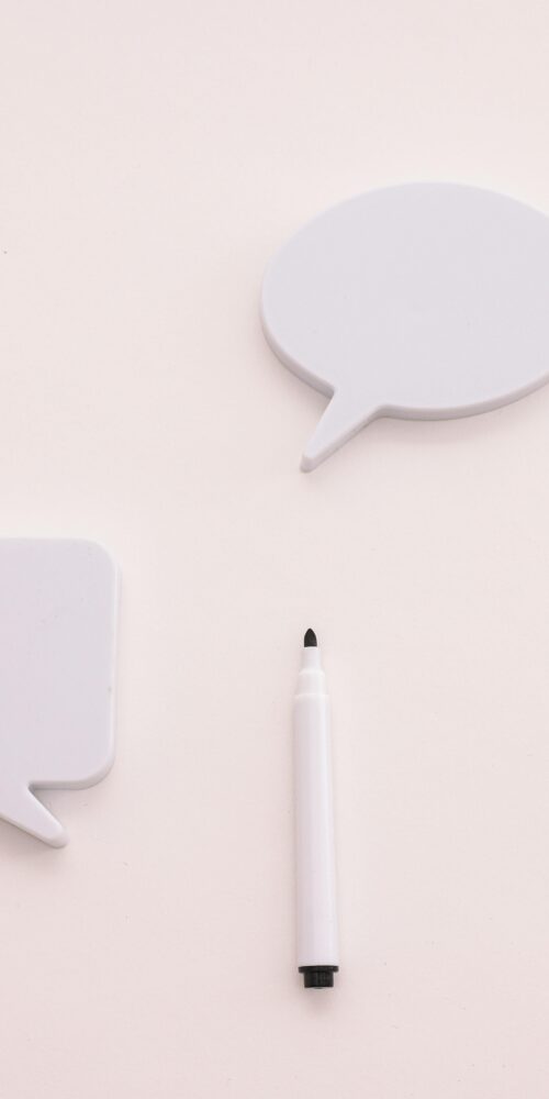 Two blank white speech bubble cutouts and a white marker placed on a light background, symbolizing communication and conversation.