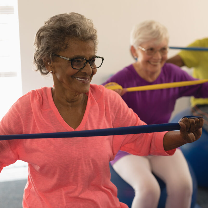 Three elderly patients sitting on exercise balls and using resistance bands for geriatric physical therapy exercises.
