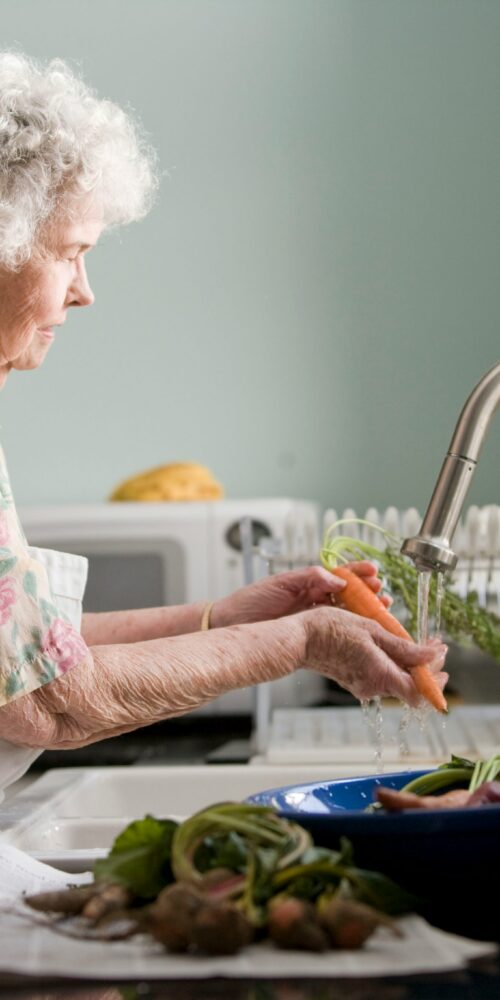 A senior woman stands at the kitchen sink and washes produce to signify the daily activities someone with a cognitive communication deficit may struggle with.