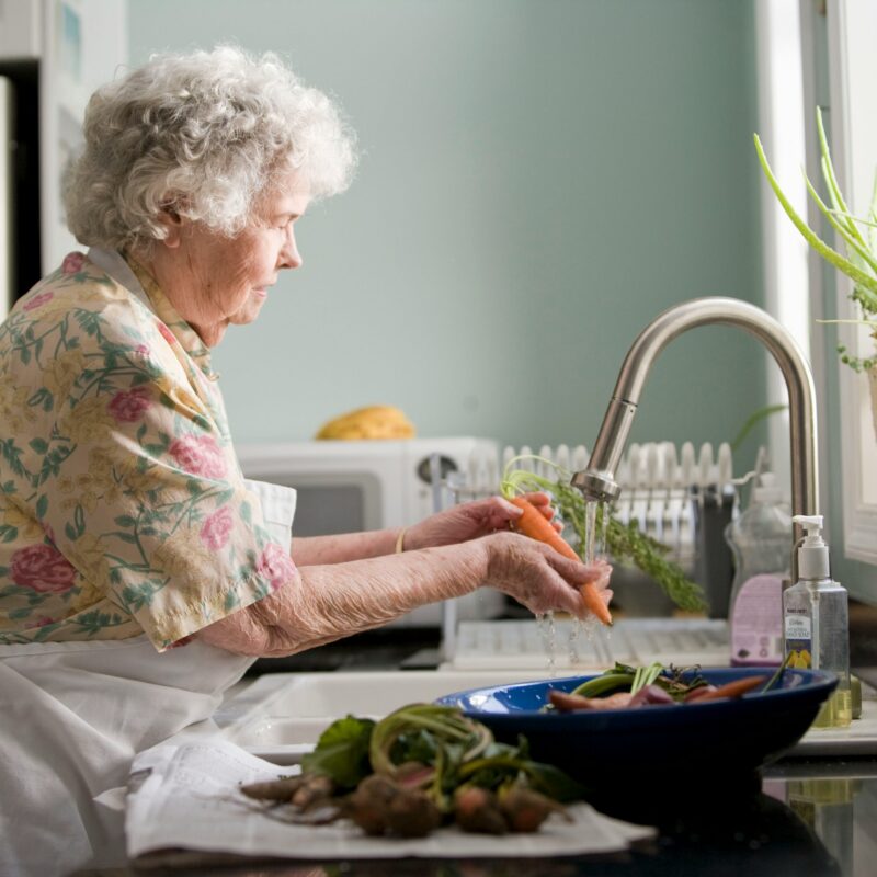A senior woman stands at the kitchen sink and washes produce to signify the daily activities someone with a cognitive communication deficit may struggle with.