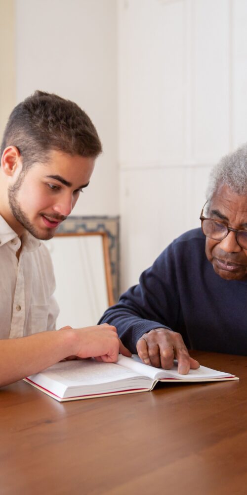 A young man sits with an older man. The older man reads from a book, practicing techniques in adult speech therapy.
