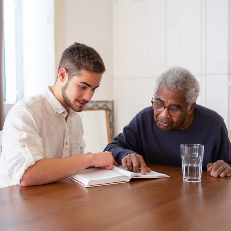 A young man sits with an older man. The older man reads from a book, practicing techniques in adult speech therapy.